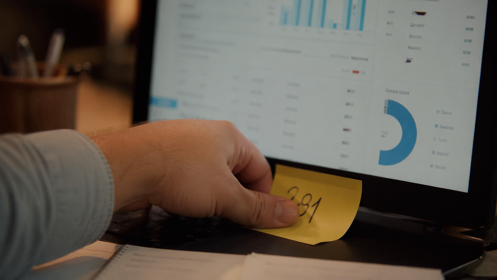 Close-up of a man's hand pressing a yellow sticky note with a single handwritten number onto the bottom edge of a laptop monitor, with a soft out-of-focus dashboard behind